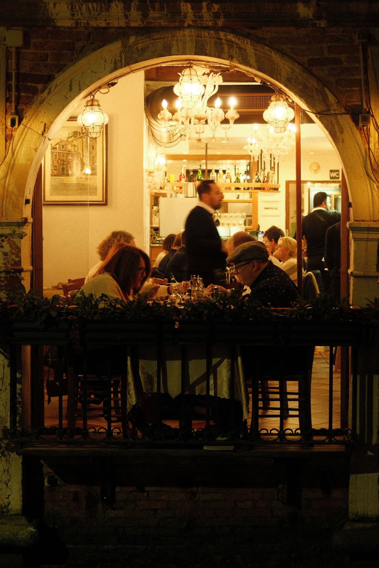 Sala di osteria illuminata a luce calda, vista di sera oltre un arco in pietra e una cancellata in ferro battuto